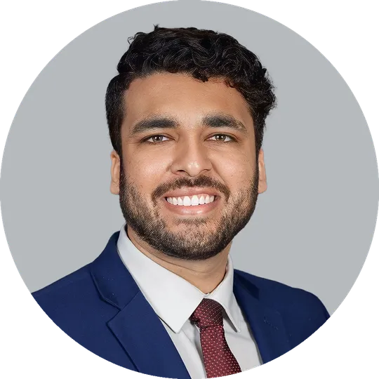 Portrait of Dr. Anuj Shah wearing a navy suit, white shirt, and burgundy tie, smiling with a light gray background