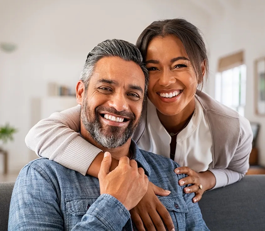 A smiling woman leaning over to hug a smiling man seated on a sofa