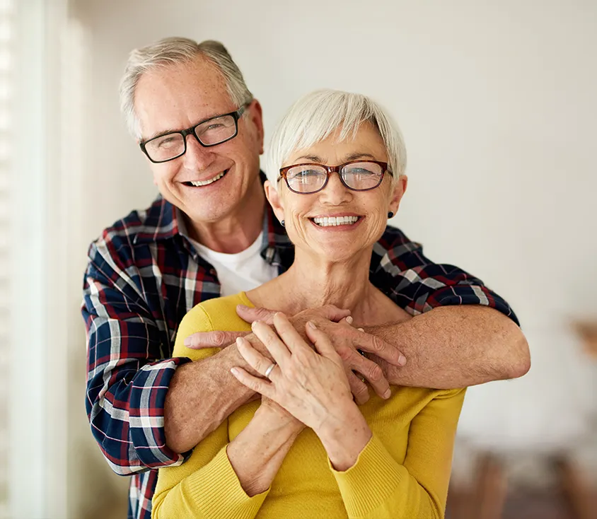 An older man in a plaid shirt hugging a smiling older woman in a yellow sweater from behind, both wearing glasses