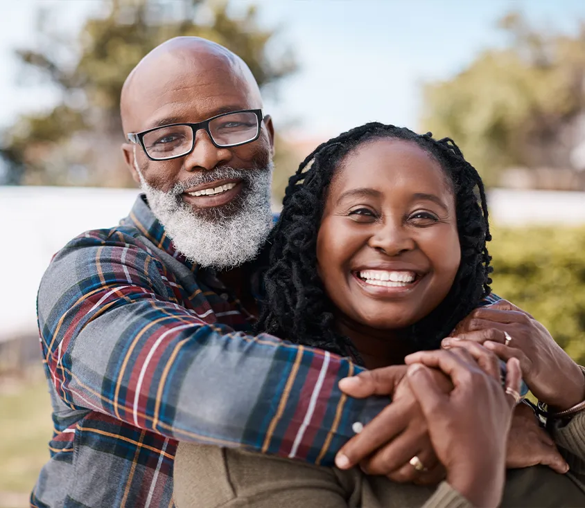 A smiling couple embracing outdoors, the man wearing glasses and a plaid shirt