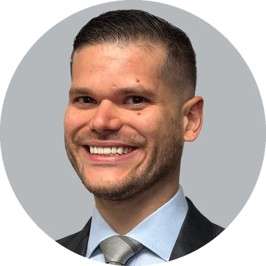Portrait of Dr. Lucas Bracero wearing a dark suit, light blue shirt, and silver tie, smiling with a light gray background