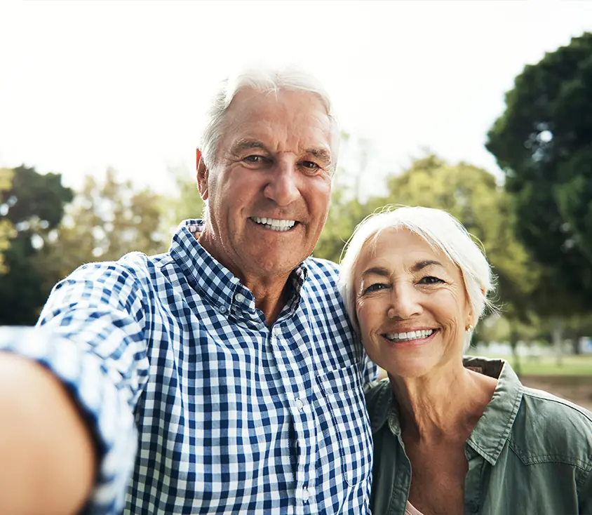 about-the-spine-and-nerve-centers-of-the-virginias-2 An older couple smiling at the camera while standing outdoors. The man is wearing a blue and white checkered shirt, and the woman is wearing a green shirt. They are standing close together, with trees and greenery in the background