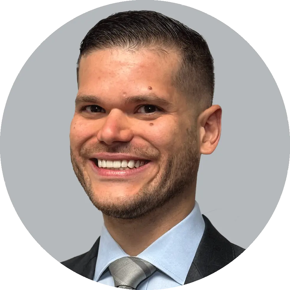 Portrait of Dr. Lucas Bracero wearing a dark suit, light blue shirt, and silver tie, smiling with a light gray background